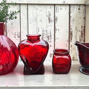 May include: A collection of four red glass decorative items. Includes a round vase, a heart-shaped vase, a small jar, and a creamer pitcher. The items are arranged on a white surface against a distressed white wooden background.