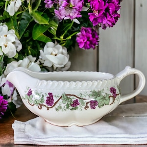 May include: White ceramic gravy boat with a grapevine design and purple grapes. The boat is sitting on a white napkin on a wooden table.