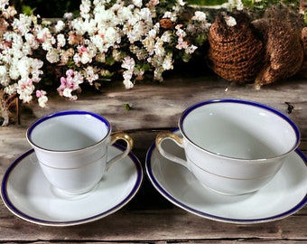 May include: Two white porcelain teacups with blue and gold trim, one larger than the other, on white saucers with blue and gold trim, on a wooden surface.