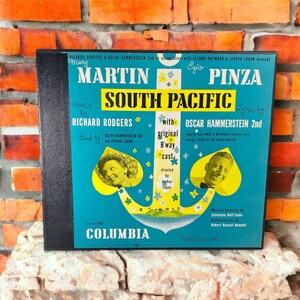 May include: A vintage Columbia Records album cover featuring Mary Martin and Ezio Pinza in the musical South Pacific. The cover is blue with a large anchor and a yellow heart shape. The title "South Pacific" is in large yellow letters. The text "Music by Richard Rodgers" and "Lyrics by Oscar Hammerstein 2nd" is also visible.