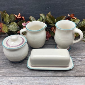 May include: A set of four white ceramic kitchenware pieces with a teal and purple trim. The set includes a creamer, a sugar bowl with lid, a butter dish, and a mug.
