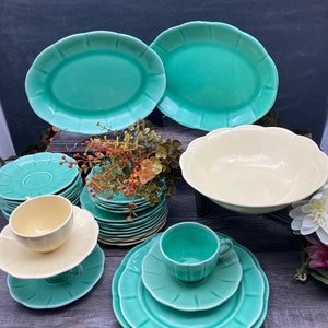 May include: A set of vintage green and cream colored ceramic dinnerware. The set includes a large bowl, two oval platters, a stack of dinner plates, a stack of salad plates, a stack of saucers, and two teacups.