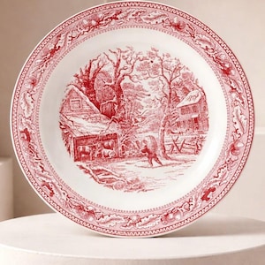 May include: A white ceramic plate with a red transferware design. The central image depicts a winter scene with a house, trees, and figures. The rim features a decorative border. A classic piece of tableware.