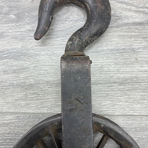May include: An antique, weathered metal pulley system with a large hook and a spoked wheel. The hook is curved and shows signs of age and use. The wheel is partially visible, with spokes radiating from the center.