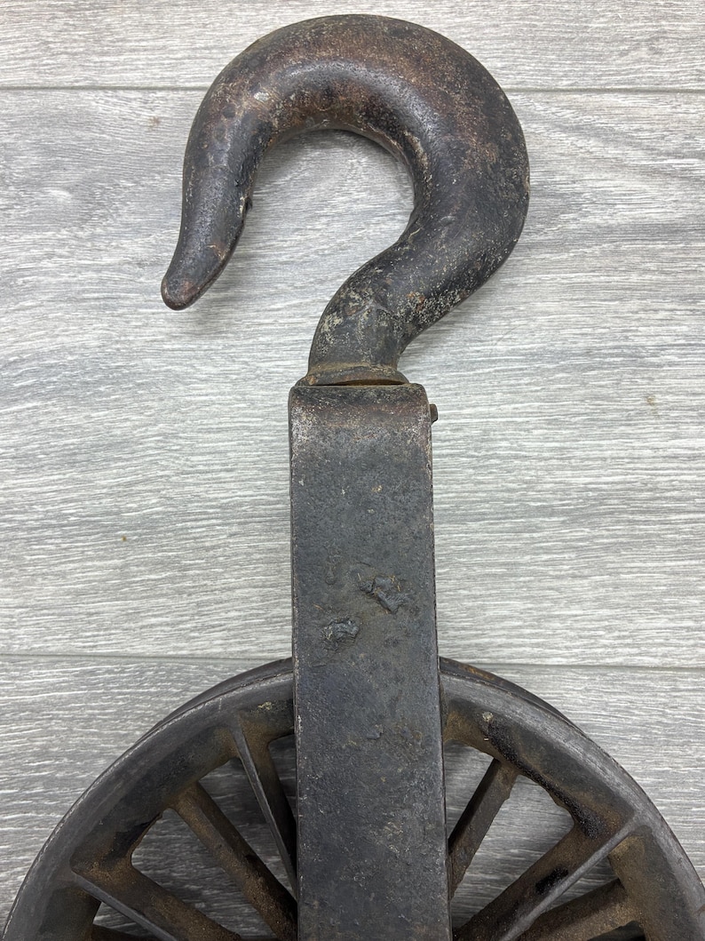 May include: An antique, weathered metal pulley system with a large hook and a spoked wheel. The hook is curved and shows signs of age and use. The wheel is partially visible, with spokes radiating from the center.