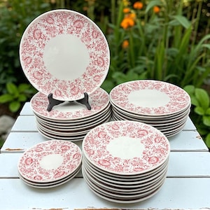 May include: A collection of white ceramic plates with a red floral pattern. The plates are stacked in multiple piles, with one plate displayed on a black stand. The plates have a white center and a red floral border. The plates are on a white wooden surface.