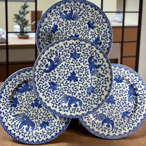 May include: A collection of five ceramic plates with a cream-colored background and intricate blue patterns. The plates feature a central design of stylized birds and floral motifs, surrounded by a detailed border. The plates are arranged in a visually appealing display.