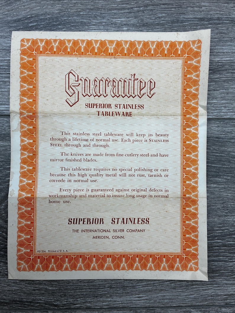 May include: A vintage guarantee card for Superior Stainless Tableware. The card features an ornate orange border and text in a classic font. The text includes a guarantee of the tableware's quality and durability, and the manufacturer's information.