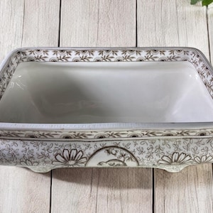 May include: A white ceramic serving dish with a floral pattern in brown and gold. The dish has a rectangular shape with a raised rim and four feet.
