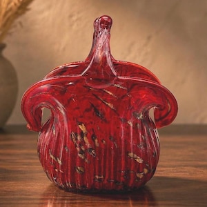 May include: A vibrant red glass basket featuring a vertical ribbed texture and scattered gold-colored flecks throughout the body. The vessel has a wide, open top with two curved handles that meet at a central point, creating a distinct silhouette.