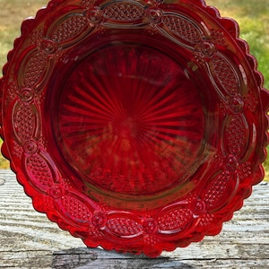 May include: A vibrant red glass plate with a scalloped edge. The plate features a detailed, embossed design around the rim and a sunburst pattern in the center. The plate is likely vintage and adds a touch of elegance.