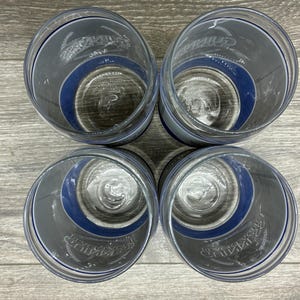 May include: Four clear drinking glasses with a dark gray and blue design. The glasses have a blue band around the middle and a logo printed on the side. The glasses are arranged in a square on a wooden surface.