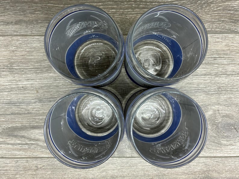 May include: Four clear drinking glasses with a dark gray and blue design. The glasses have a blue band around the middle and a logo printed on the side. The glasses are arranged in a square on a wooden surface.