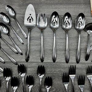 May include: A collection of stainless steel silverware, including spoons, forks, and serving utensils. The spoons have a decorative cut-out design. The silverware is arranged on a gray wooden surface.