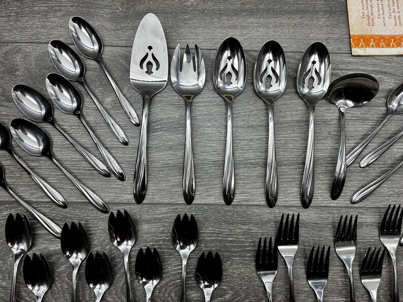 May include: A collection of stainless steel silverware, including spoons, forks, and serving utensils. The spoons have a decorative cut-out design. The silverware is arranged on a gray wooden surface.