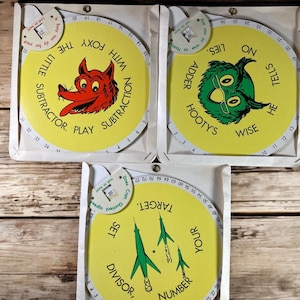 May include: Three colorful educational game wheels with yellow backgrounds and cartoon characters. The first wheel features a red fox with the text "The Foxy Little Subtractor. Play with Subtraction". The second wheel features a green owl with the text "Wise He Tells On Lies. Adder Hooty's". The third wheel features three green rockets with the text "Your Number Target. Set Divisor".