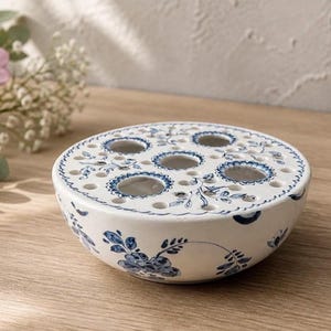 May include: A round white ceramic flower frog featuring a blue floral pattern. The top surface has multiple circular openings of varying sizes designed for arranging stems, with a smooth, curved bowl-shaped base decorated with painted blue flowers.