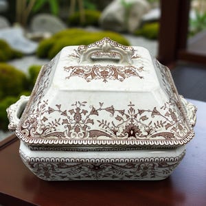 May include: A white and brown antique serving dish with a lid. The dish has a square shape with rounded corners and a decorative handle on top. The dish is decorated with a brown floral pattern.