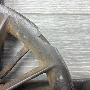 May include: Close-up of a weathered, dark metal wheel rim. The rim has a spoke design with triangular openings. The metal shows signs of rust and age, with a textured, aged appearance. The background is a gray wooden surface.