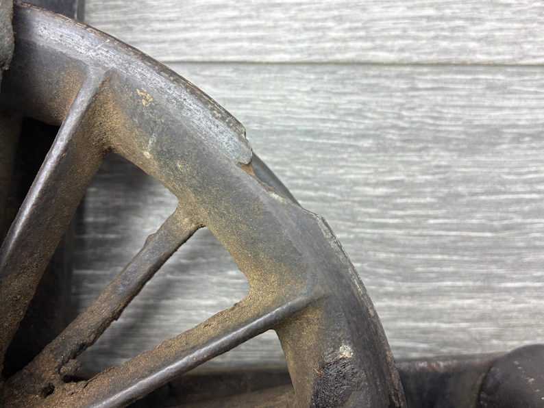 May include: Close-up of a weathered, dark metal wheel rim. The rim has a spoke design with triangular openings. The metal shows signs of rust and age, with a textured, aged appearance. The background is a gray wooden surface.