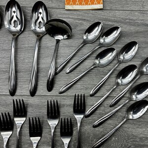 May include: A collection of stainless steel silverware, including spoons, forks, and a ladle, arranged on a gray wooden surface. The spoons have a decorative design on the handles. The forks have four tines.