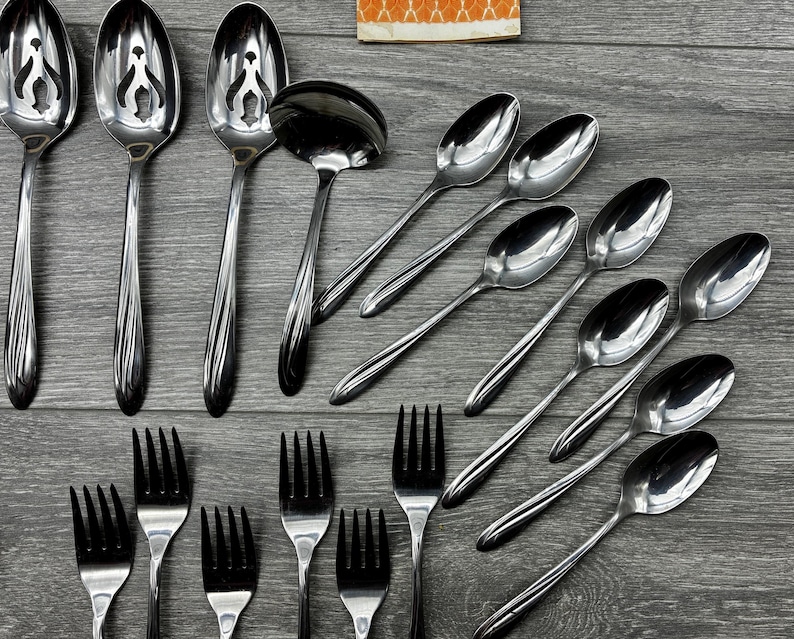 May include: A collection of stainless steel silverware, including spoons, forks, and a ladle, arranged on a gray wooden surface. The spoons have a decorative design on the handles. The forks have four tines.