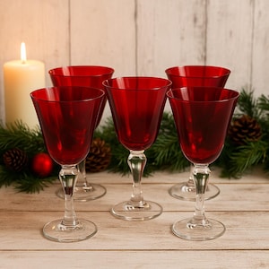 May include: Five red wine glasses with clear stems and bases are arranged on a wooden surface. A lit candle and evergreen garland with pinecones and red ornaments are in the background. The glasses have a classic, elegant design.