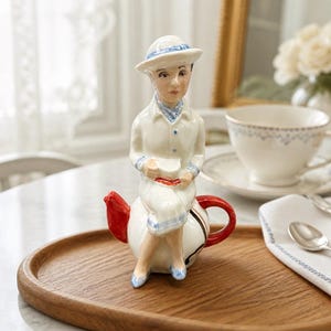 May include: A ceramic teapot shaped like a person sitting down, wearing a white outfit with blue accents and a matching hat. The teapot features a bright red spout and a red handle, with the figure holding a small white cup in their hands.