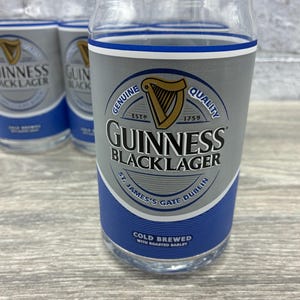May include: Clear glass tumblers with a design that mimics a Guinness Black Lager can. The design includes the Guinness logo, a harp, and text that reads "Genuine Quality" and "St. James's Gate Dublin". The tumblers are on a light wood surface.