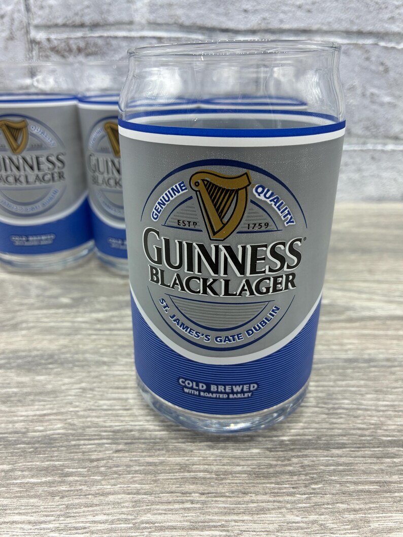 May include: Clear glass tumblers with a design that mimics a Guinness Black Lager can. The design includes the Guinness logo, a harp, and text that reads "Genuine Quality" and "St. James's Gate Dublin". The tumblers are on a light wood surface.