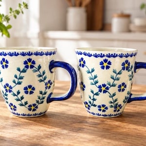 May include: Two ceramic mugs with a cream-colored base featuring a blue and yellow floral pattern arranged in a diamond lattice design. Each mug has a solid blue handle and a blue scalloped border along the top rim of the vessel.