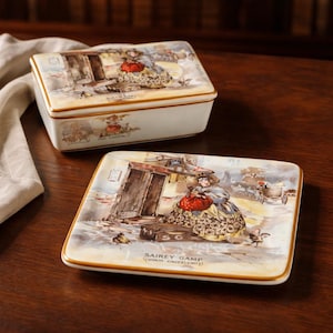May include: A rectangular trinket box and a square plate with a vintage illustration. The illustration depicts a woman in a large dress with a red bag, standing near a doorway. The plate has the text "SAIREY GAMP (MAKIN' CRUMZLEWITS)".