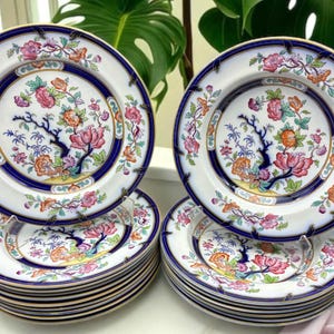May include: A collection of vintage porcelain plates. Each plate features a white background with a blue border and a floral design in pink, orange, and green. The plates are stacked in sets, showcasing their intricate patterns and colors.