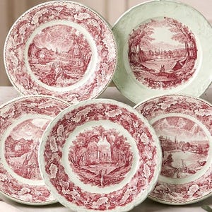 May include: A collection of decorative plates with a red and white landscape design. The plates feature a detailed illustration of a scenic view, surrounded by a decorative border. The plates are arranged on a light-colored surface.