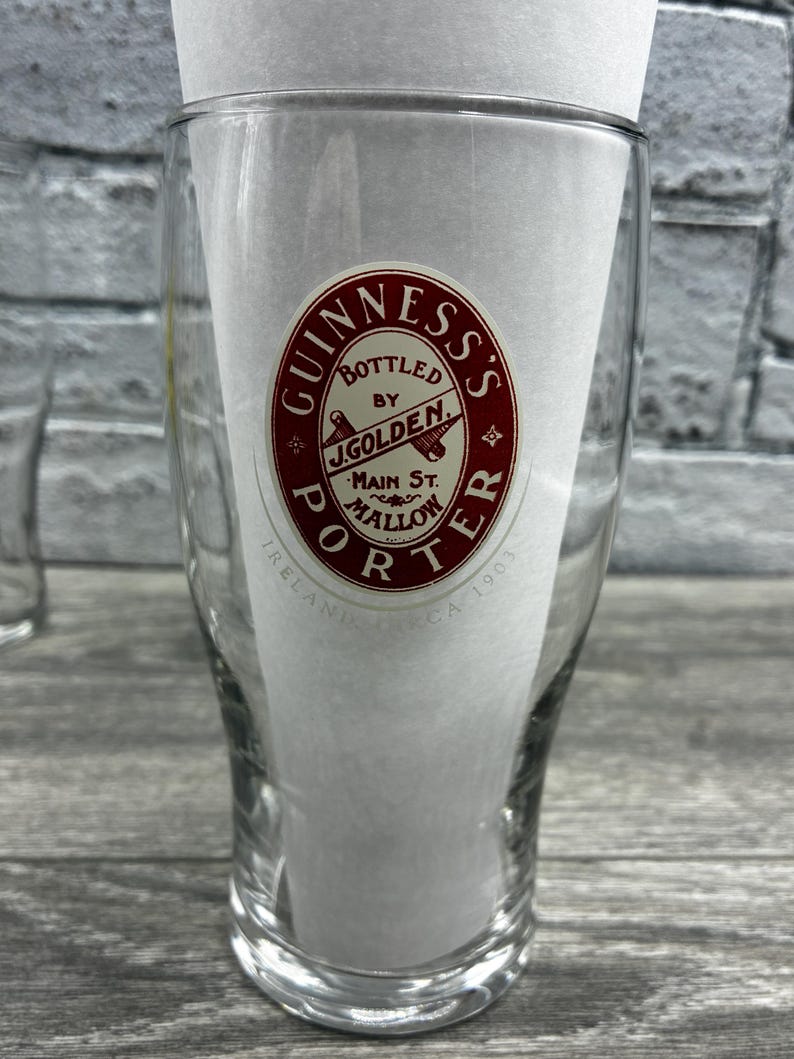 K&ouml;nnte beinhalten: Klares Pintglas mit dem Guinness's Porter Logo in Rot und Wei&szlig;. Das Logo enth&auml;lt den Text "Bottled by J. Golden, Main St. Mallow, Ireland, CA 1903". Das Glas ist teilweise mit wei&szlig;em Papier gef&uuml;llt.