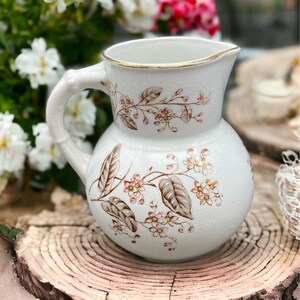 May include: A white ceramic pitcher with a floral design. The pitcher has a round body and a handle. The floral design features brown leaves and pink flowers.