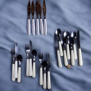 May include: A set of white-handled silverware, including knives, forks, and spoons. The knives have a gold-colored metal blade and the forks and spoons have a silver-colored metal head. The handles are all white with a subtle pattern.