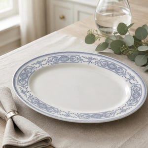 May include: An oval white serving platter with a blue decorative border. The border features a repeating pattern of floral and geometric designs. A folded linen napkin and a silver napkin ring are in the foreground.