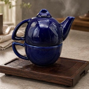 May include: A cobalt blue ceramic teapot and teacup set. The teapot sits atop the teacup, with steam rising from the spout. The set rests on a dark wooden tray, creating a striking contrast.
