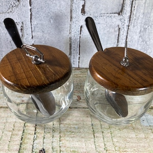 May include: Two glass jars with wooden lids and spoons inside. The lids are dark brown wood with a metal ring and a spoon attached to the lid.