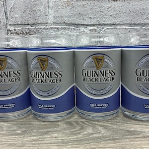 May include: Four clear glass tumblers with a silver and blue design. Each glass features the Guinness logo and the words "Guinness Black Lager". The glasses are cold brewed and have a capacity of approximately 16 ounces.