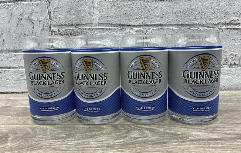 May include: Four clear glass tumblers with a silver and blue design. Each glass features the Guinness logo and the words "Guinness Black Lager". The glasses are cold brewed and have a capacity of approximately 16 ounces.