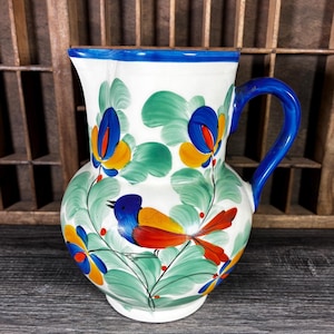 May include: White ceramic pitcher with a blue handle and rim, adorned with a colorful bird and floral design. The bird features blue, orange, and red hues, while the flowers have blue, yellow, and green accents. A decorative piece for the kitchen.