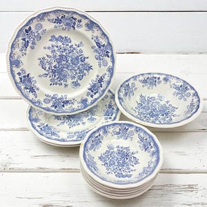 May include: A collection of white ceramic dishes with blue floral patterns. The set includes plates and bowls with scalloped edges. The floral design is centered on each piece and repeated around the rims. The dishes are arranged on a white wooden surface.