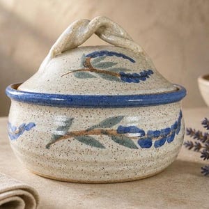 May include: A round ceramic lidded jar with a speckled cream finish, featuring hand-painted blue and green lavender sprigs. The lid has a twisted handle, and the rim of the jar is accented with a solid blue band, creating a cohesive design.