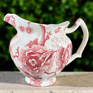 May include: A white ceramic pitcher with a red floral pattern. The pitcher has a curved spout and a handle. The floral design includes large flowers and smaller floral accents. The pitcher is on a stone surface.