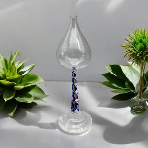 May include: A clear glass decorative piece with a teardrop-shaped top and a round base. The stem is adorned with a twisted pattern of blue and gold. Green plants flank the glass object.
