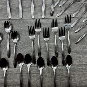 May include: An assortment of stainless steel silverware, including forks, spoons, and serving utensils, arranged on a gray wooden surface. The silverware has a reflective, polished finish, and the arrangement suggests a formal dining setting.