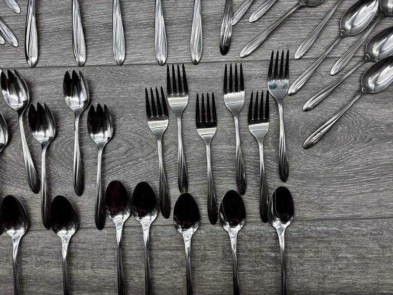 May include: An assortment of stainless steel silverware, including forks, spoons, and serving utensils, arranged on a gray wooden surface. The silverware has a reflective, polished finish, and the arrangement suggests a formal dining setting.