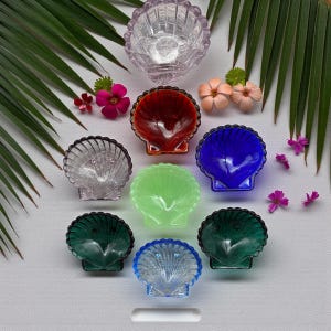 May include: A collection of colorful, seashell-shaped glass dishes. The dishes are in various colors, including clear, red, blue, green, and purple. The image also includes decorative flowers and palm leaves.
